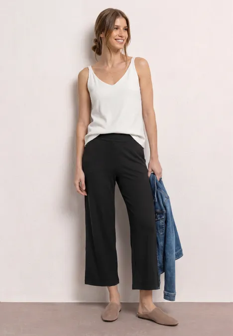 Vrouw in wit topje, zwarte culottes en taupekleurige mules, met een spijkerjasje in de hand.