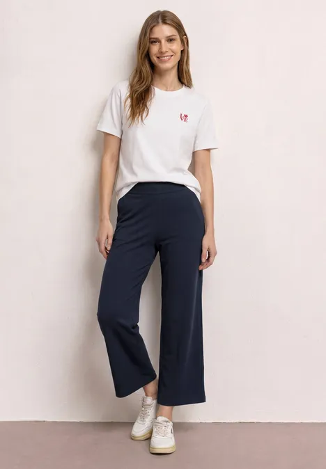 Vrouw voor beige achtergrond, draagt een wit t-shirt, wijde marineblauwe broek en witte sneakers.