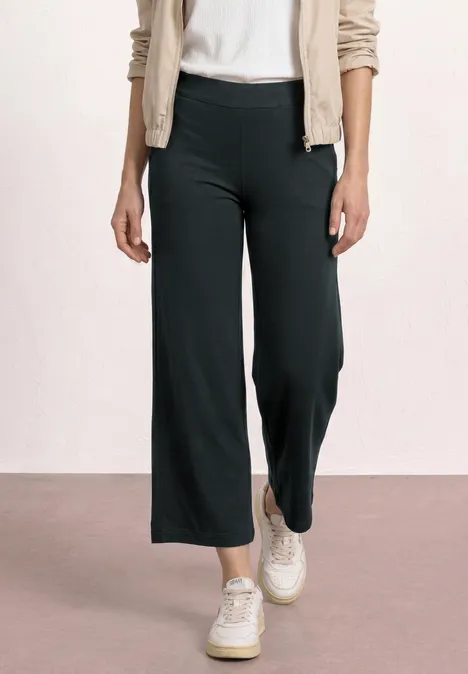 Vrouw in zwarte wijde 7/8-broek met witte sneakers en beige vest