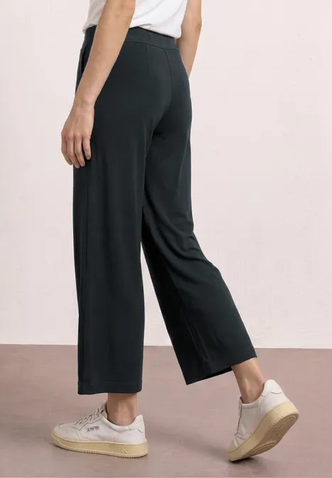 Achteraanzicht van een persoon in zwarte, wijdvallende 3/4-broek en witte sneakers.