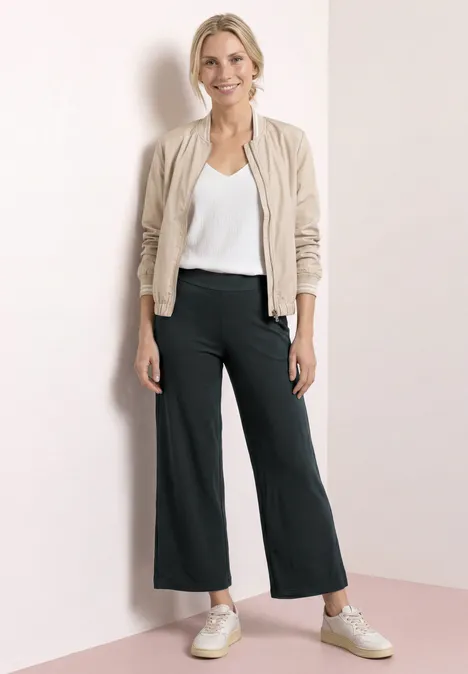 Vrouw in zwarte culotte, wit topje, beige bomberjack en sneakers voor neutrale achtergrond.
