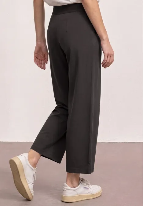 Vrouw met zwarte, wijde broek en witte sneakers, zijaanzicht tegen neutrale achtergrond.