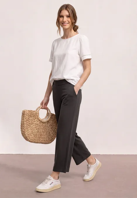 Vrouw in wit T-shirt, zwarte broek, beige sneakers, met een strooien tas met ronde handvatten.