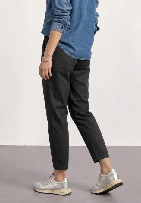 Zijaanzicht van persoon in donkergrijze broek en zilverkleurige sneakers voor een witte muur.