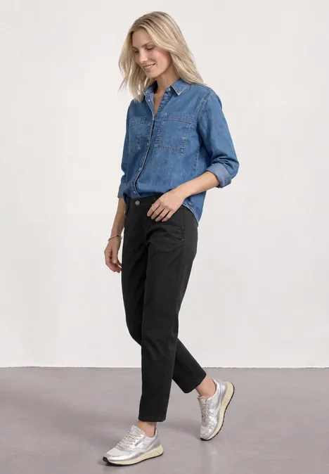Vrouw in denim blouse, zwarte broek en witte sneakers voor een neutrale achtergrond.