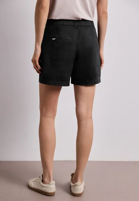 Person von hinten in schwarzen Shorts, weiße Sneakers, neutrale Wand im Hintergrund