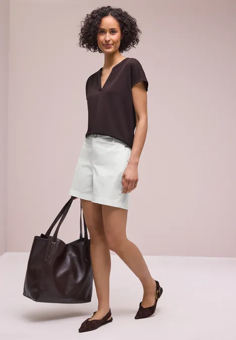 Vrouw met krullend haar draagt een donker T-shirt, lichte rok, zwarte ballerina’s en een grote bruine tas.