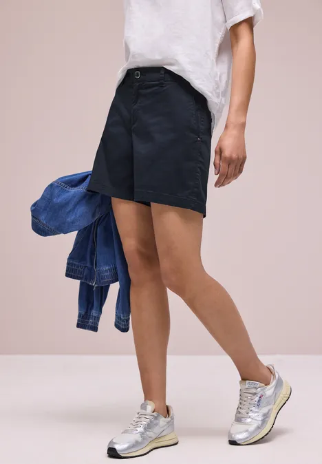 Eine Person trägt dunkelblaue Shorts, weißes T-Shirt, hält eine Jeansjacke und trägt silberne Sneaker.