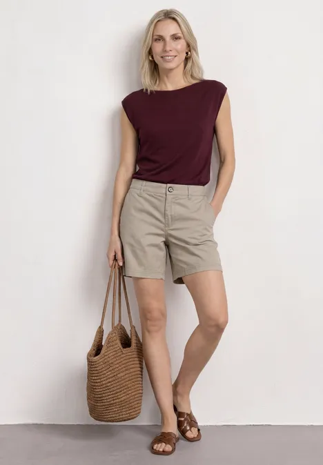 Frau in bordeauxrotem Oberteil, beigen Shorts und braunen Sandalen mit Strohtasche vor hellem Hintergrund.
