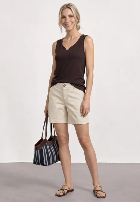 Frau in braunem Top und beigen Shorts mit Sandalen und gestreifter Tasche vor weißem Hintergrund.
