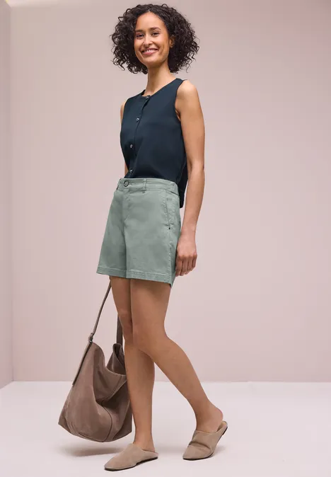 Vrouw met krullend haar in een mouwloos marineblauw topje, lichtgroen rokje, beige mules en een grote tas.