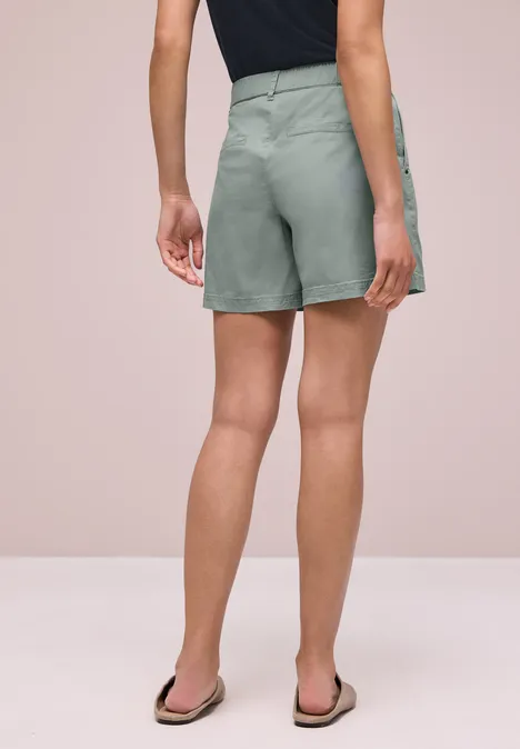 Vrouw van achteren in lichtgroene, hooggetailleerde short en slippers voor een beige achtergrond.