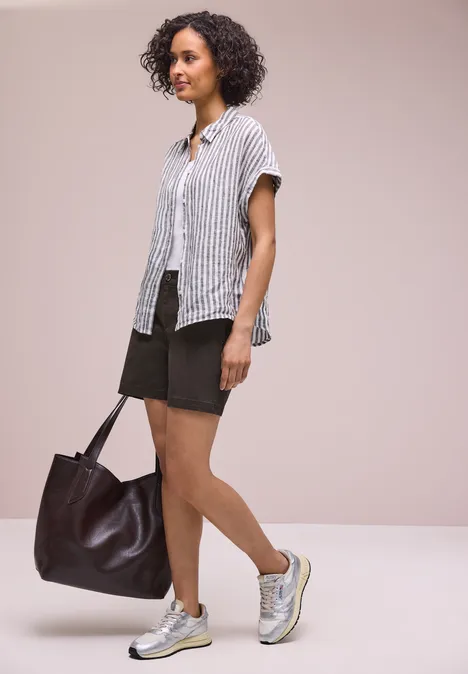 Vrouw met krullend haar draagt gestreept overhemd, zwarte shorts, sneakers en een grote bruine tas.