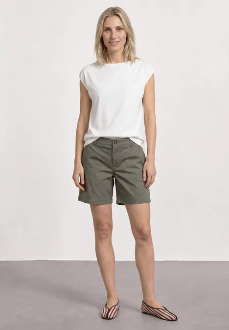 Eine Frau in weißem T-Shirt, khakigrünen Shorts und gestreiften Pantoletten vor neutralem Hintergrund.