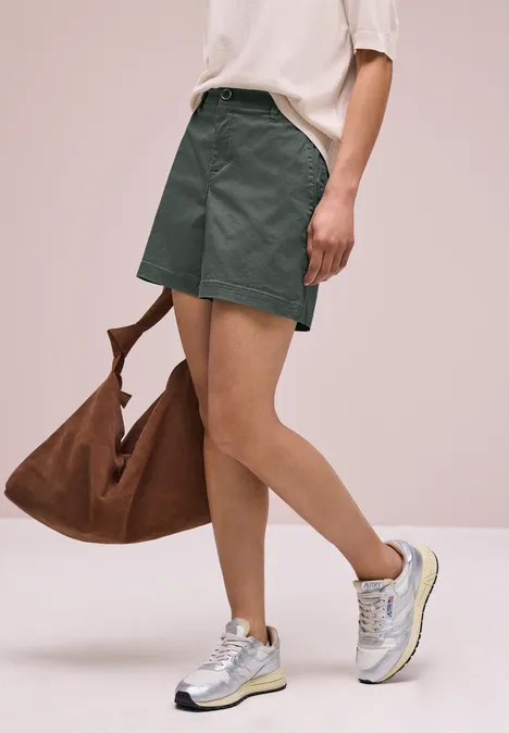 Unterkörper einer Person in grünen Shorts, weißen Sneakern und brauner Tasche.