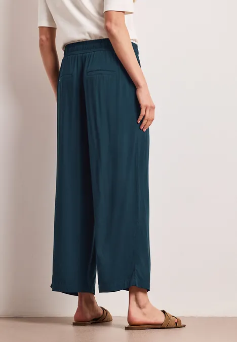 Person trägt weite, knöchellange, petrolfarbene Hose und Sandalen, Rückansicht.