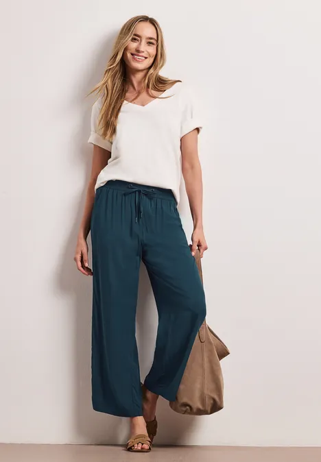 Frau in weißem T-Shirt und weiten, teal-farbenen Hosen trägt Sandalen und Tasche