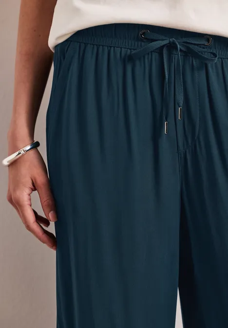 Frau trägt weite, dunkelblaue Bundhose mit Kordelzug, weiße Bluse und Armbänder am Handgelenk.