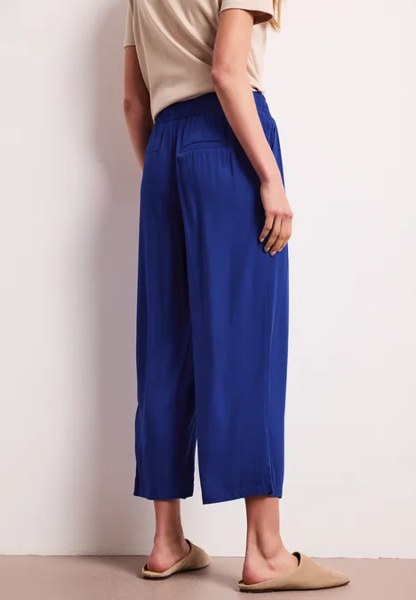 Eine Frau trägt ein beiges Oberteil mit leuchtend blauen, weiten 7/8-Hosen und beigen Pantoletten.