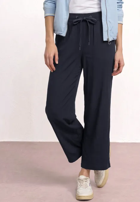 Eine Person trägt dunkelblaue, weite Hosen, weiße Sneaker und eine leichte Jacke.