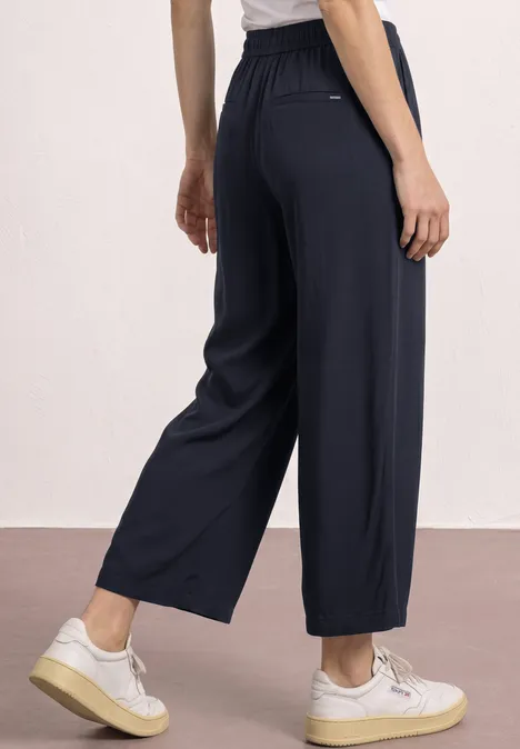 Frau in weiten, knöchellangen, dunkelblauen Hosen mit hohem Bund und weißen Sneakern.