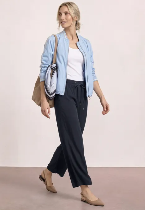 Frau in hellblauer Jacke, weißem Top, dunkler Hose und Nude-Ballerinas mit zwei Taschen.