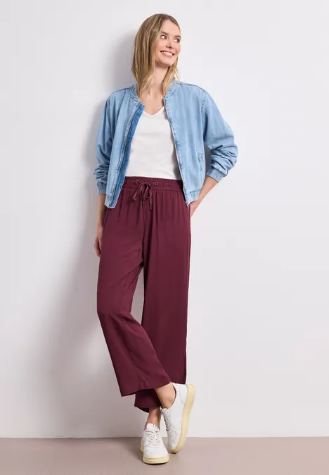 Frau lehnt an weißer Wand und trägt weißes Shirt, helle Jeansjacke, weinrote weite Hose und weiße Sneaker.