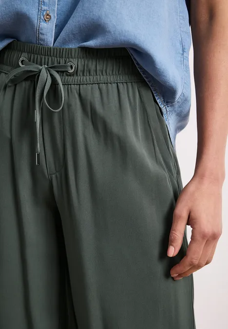 Person trägt olivgrüne Jogginghose mit Tunnelzug und hellblaues Hemd, rechte Hand an der Seite.