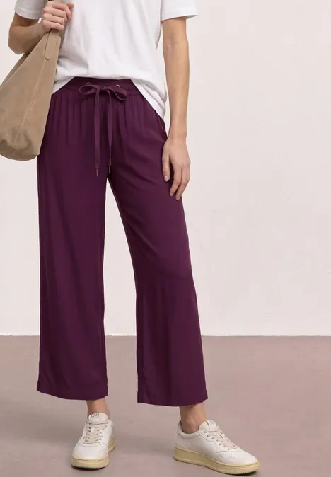 Person trägt weite, lila Hose, weißes T-Shirt, weiße Sneaker und hält eine beige Stofftasche.