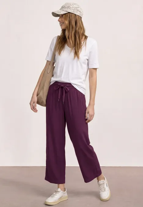 Frau mit weißem T-Shirt, lila weiten Hosen, Sneakern und Basecap steht vor schlichter Wand.