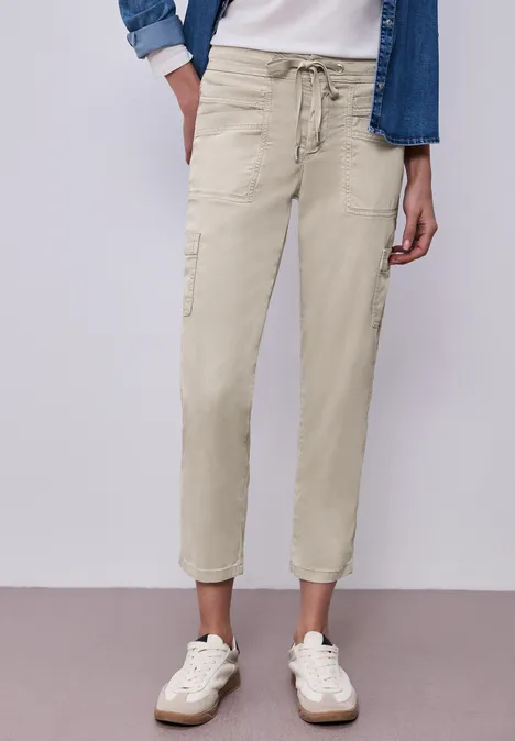 Beige Cargo-Hose mit Tunnelzug, seitlichen Taschen und weißen Sneakern an neutraler Wand.