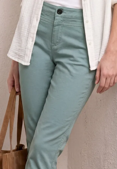 Draagt een witte blouse, een open textuurovershirt, mintgroene chino en houdt een bruine tas vast.
