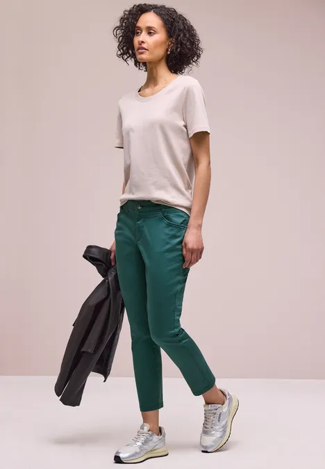 Frau trägt weißes T-Shirt, grüne Hose und Sneaker, hält eine dunkle Jacke.