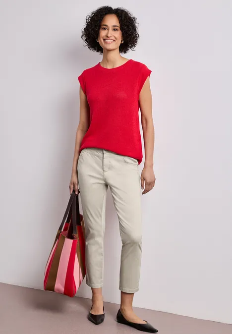 Vrouw in rood hemd, beige broek en gestreepte tas, zwarte schoenen, neutrale achtergrond.