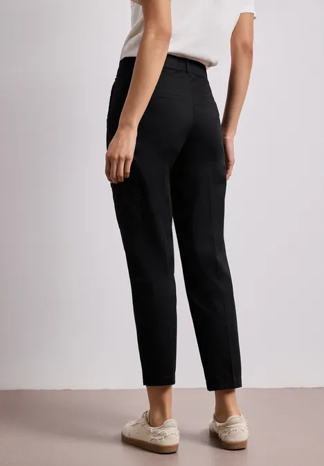 Persoon van achteren in zwarte high-waist broek en beige sneakers tegen neutrale achtergrond.