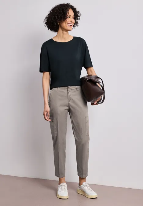 Vrouw met zwart T-shirt, grijze smalle broek, witte sneakers en een bruine tas.