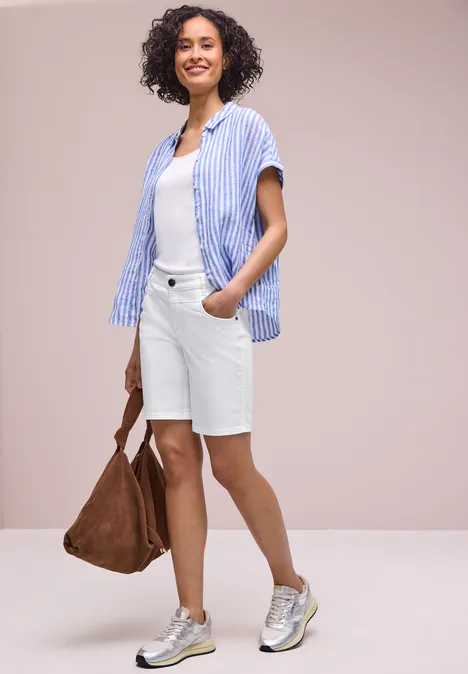 Vrouw in een casual zomeroutfit: gestreept overhemd, wit T-shirt, witte bermuda, zilverkleurige sneakers en bruine tas.