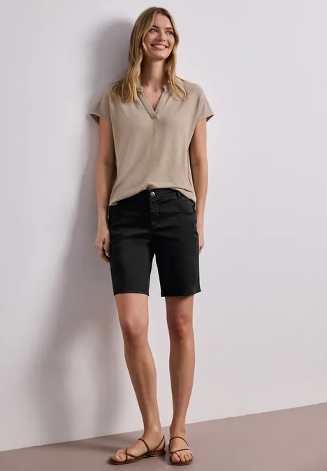 Vrouw in beige top, zwarte jeansshort en sandalen voor een effen achtergrond.