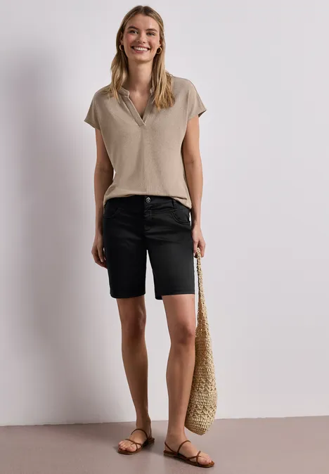 Vrouw met beige T-shirt, zwarte bermudashorts, draagt gevlochten tas en platte sandalen voor een lichte achtergrond.