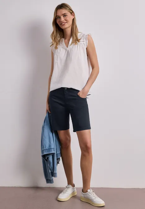 Frau trägt weiße Bluse, dunkle Shorts und weiße Sneakers, hält blaue Jeansjacke lässig in der Hand.