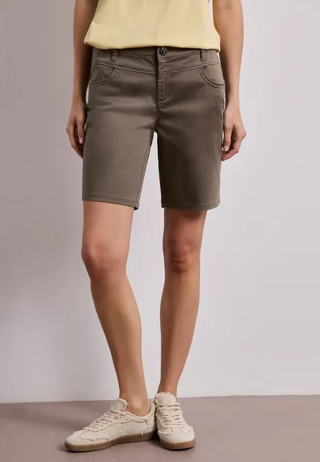 Vrouw in kaki bermudashort en witte sneakers tegen neutrale achtergrond.