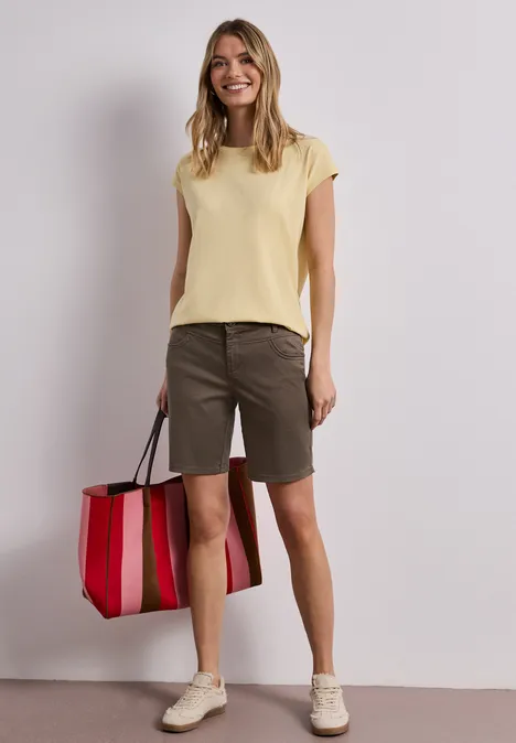 Vrouw in geel topje, groene bermuda en witte sneakers met een rood-roze gestreepte tas.