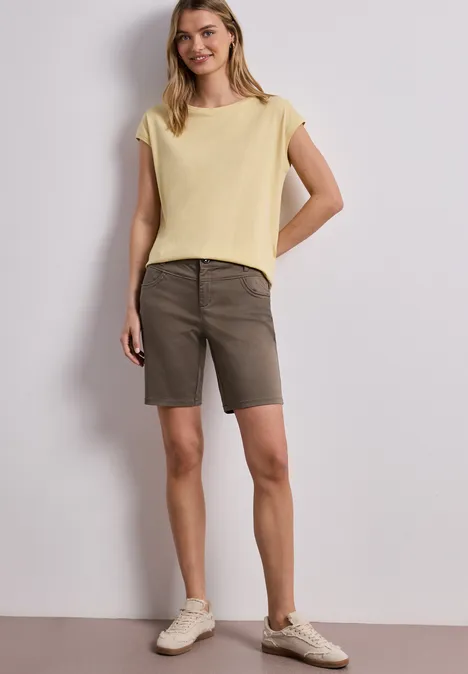 Vrouw met geel T-shirt, olijfgroene bermudashorts en beige sneakers voor een witte achtergrond.
