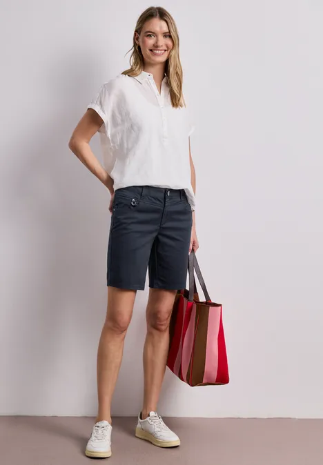 Vrouw in wit shirt, donkerblauwe bermudashort en witte sneakers met een roze-rode gestreepte shopper.