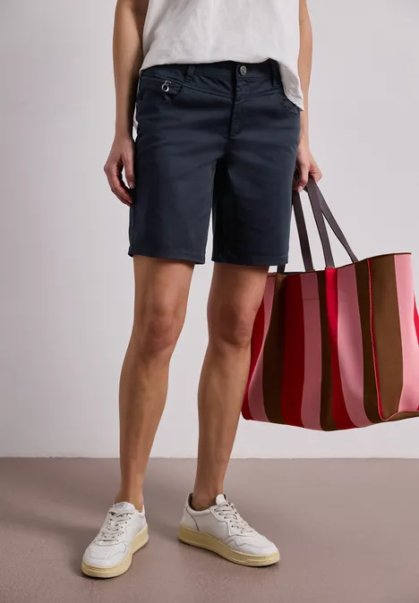 Persoon draagt marineblauwe bermudashorts, witte sneakers en houdt een gestreepte roze-rode tas vast.