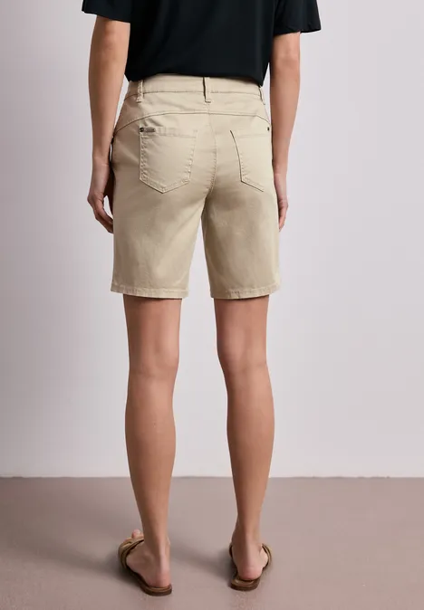 Achteraanzicht van persoon in beige bermudashort, zwart t-shirt en sandalen tegen neutrale achtergrond.
