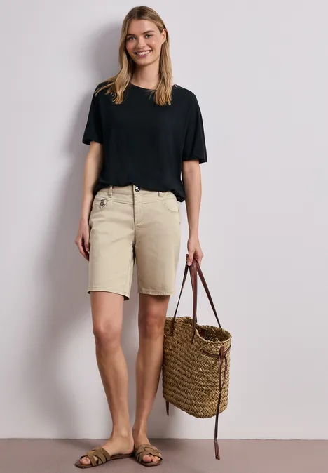 Een vrouw draagt een zwart T-shirt, beige korte broek tot op de knieën, sandalen en houdt een gevlochten rieten tas.