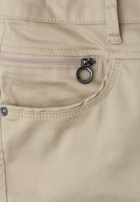 Close-up van een beige broek met een ritssluitingvak en metalen ring aan de zak.