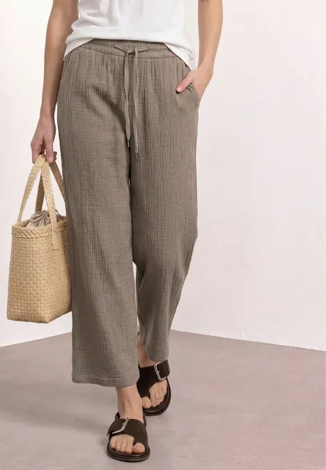 Lockere khakigrüne Leinenhose, weißes Top, schwarze Sandalen und Korbtasche.