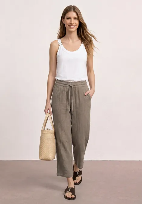 Frau trägt weißen Tanktop, lockere braune Hose, Sandalen und Strohtasche, steht vor neutralem Hintergrund.